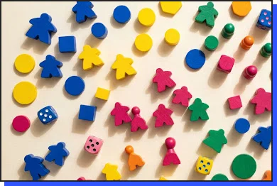 Bovenaanzicht van kleurrijke houten bordspelstukken, waaronder meepels, dobbelstenen, fiches en pionnen in verschillende kleuren zoals blauw, geel, groen en roze, verspreid over een gestreepte pastelachtergrond.